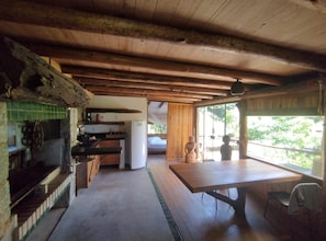 A rustic cabin interior with a wooden ceiling, a kitchen area with a refrigerator, a dining table, and a view through a large window.