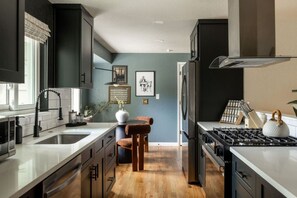 A modern kitchen with a central island, stainless steel appliances, and a dining area with a wooden chair. - Denver Dream Retreat w/Hot Tub, Sauna, Movie & Game Rms, Putting Green, Fire Pit (Arvada)