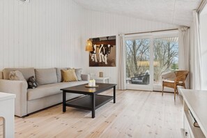 A living room with a sofa, a coffee table, and a wicker chair. There is a large window with a view of trees and a fence. - Architect-designed luxury summerhouse in Ordrup (Fårevejle)