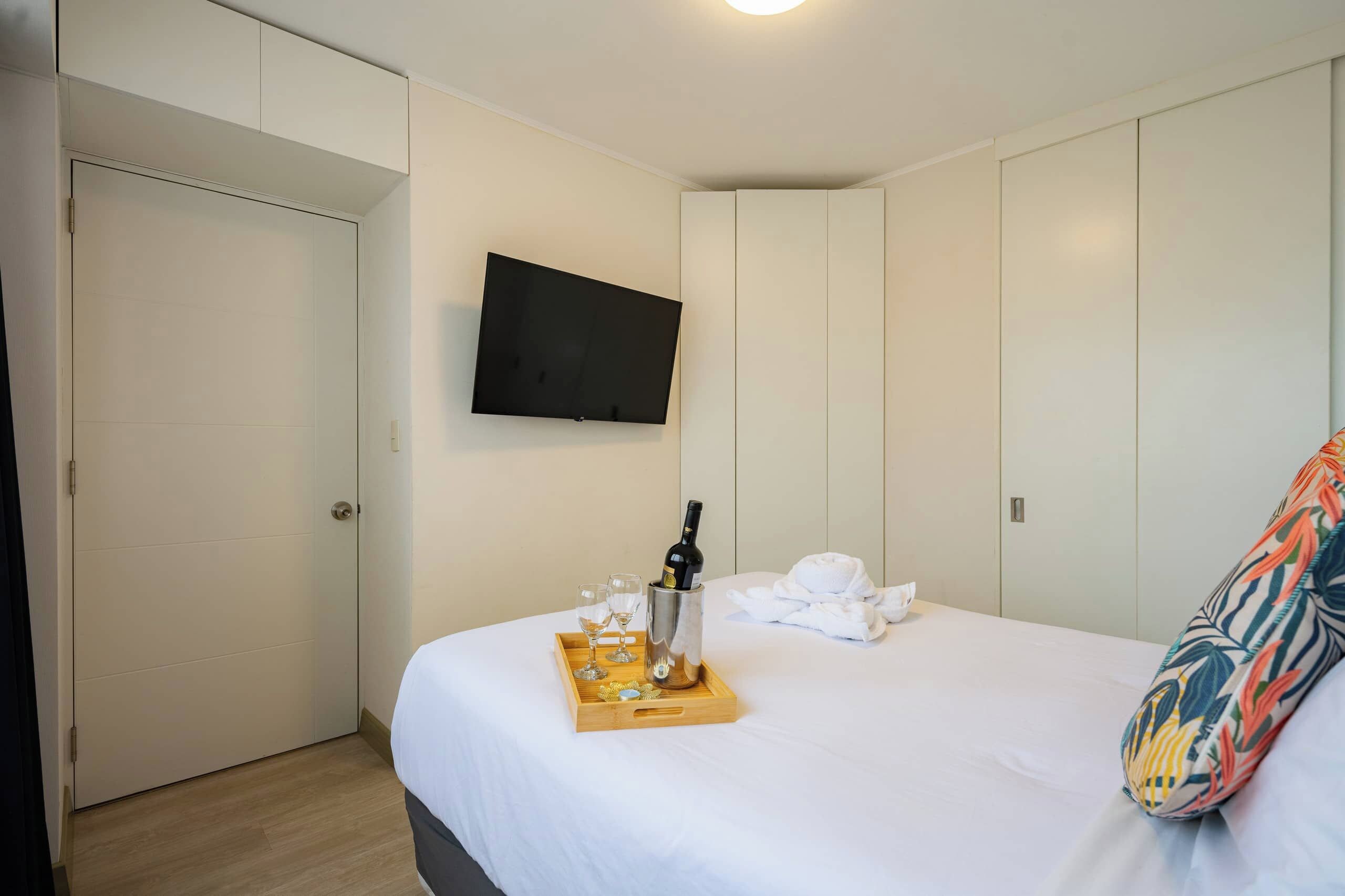 Habitación de hotel con una cama, un televisor de pantalla plana montado en la pared y una bandeja con una botella y vasos sobre la cama.