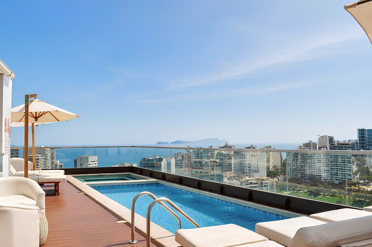 Piscina en la azotea con sillones de descanso y barandilla de cristal con vistas a la ciudad y al mar.