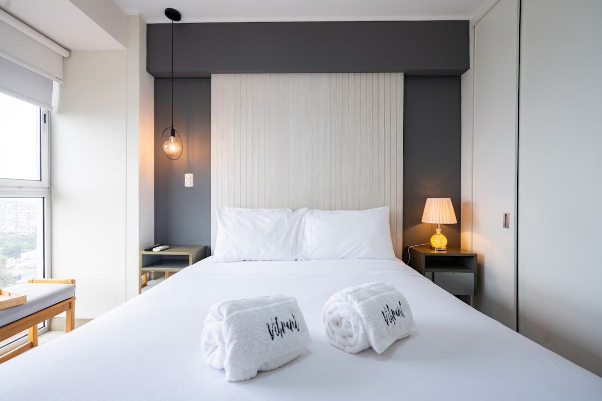Una habitación de hotel con una cama, dos toallas enrolladas, una lámpara de noche y una ventana.