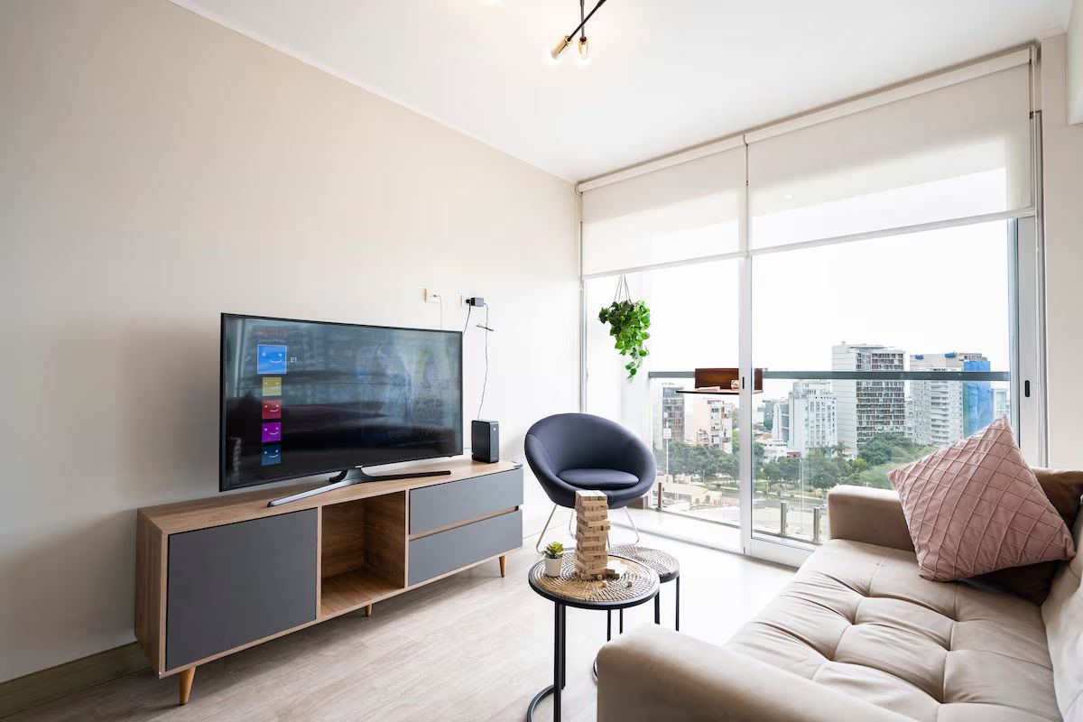 Un salon moderne doté d’un téléviseur à écran plat, d’un canapé, d’un fauteuil et d’une grande fenêtre offrant une vue sur la ville.
