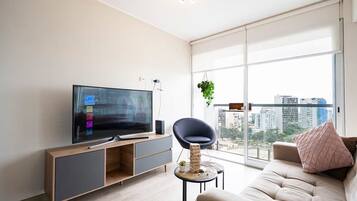 Un salon moderne doté d’un téléviseur à écran plat, d’un canapé, d’un fauteuil et d’une grande fenêtre offrant une vue sur la ville.