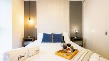 Une chambre d’hôtel avec un lit, une table de chevet, une petite commode, une lampe, un plateau avec une bouteille de vin, des verres et une petite plante.