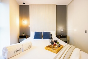 A hotel room with a bed, a bedside table, a nightstand, a lamp, a tray with a bottle of wine, glasses, and a small plant. - Connect Apartments by Vibrant (Barranco)