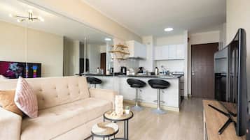 Una sala de estar moderna con un sofá, una mesa de centro y un televisor. Hay una zona de cocina con una barra y taburetes.