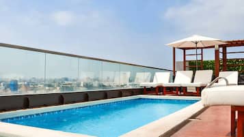 A rooftop pool with clear glass railings, white lounge chairs, and a wooden pergola with an umbrella.