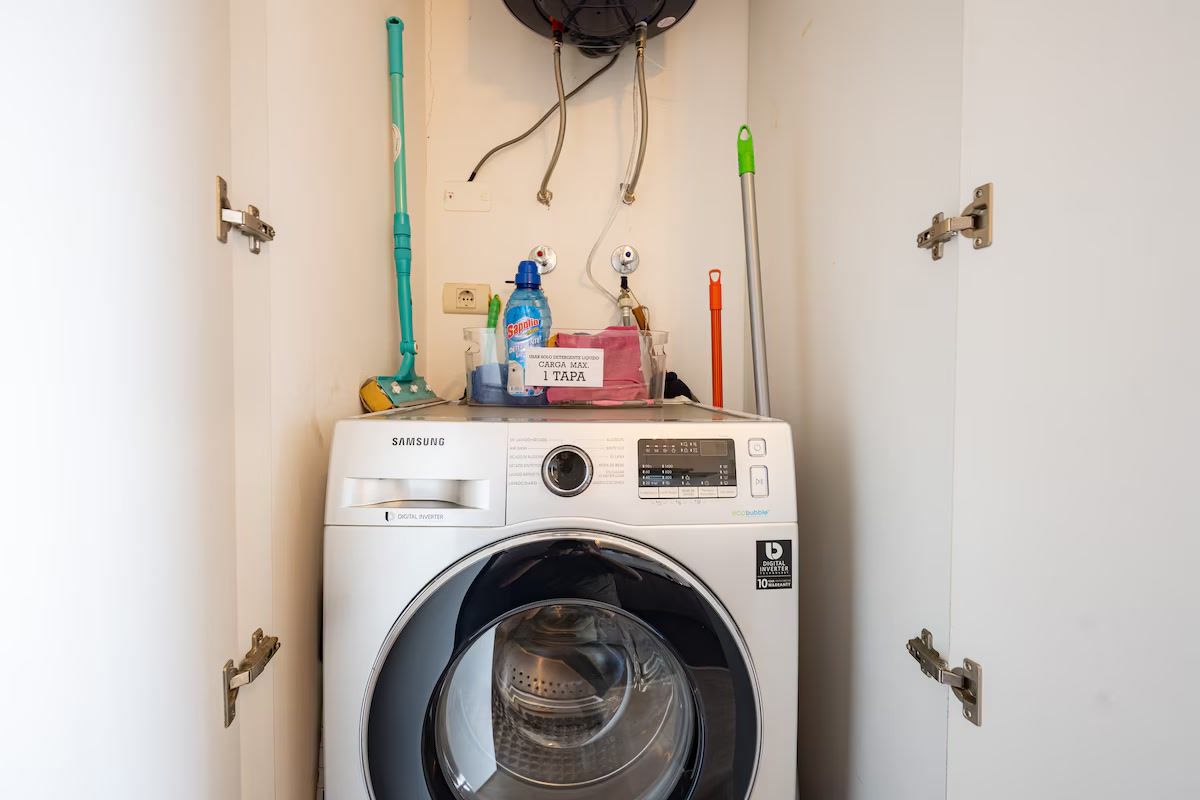 A Samsung washing machine in a utility room with a wall-mounted faucet, a mop, and a cleaning spray bottle.
