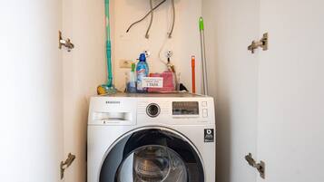 A Samsung washing machine in a utility room with a wall-mounted faucet, a mop, and a cleaning spray bottle.