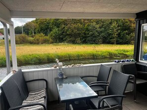 A covered outdoor seating area with a table and chairs, overlooking a river and a grassy field. - Cozy cottage by the Trend River (Løgstør)