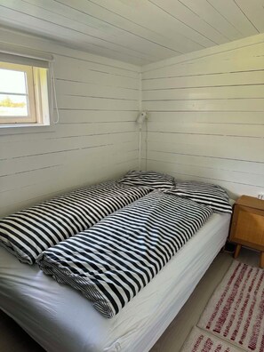 A single bed with striped bedding, a window with blinds, a wall socket, and a wooden nightstand with a lamp. - Cozy cottage by the Trend River (Løgstør)