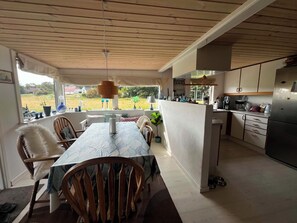 A dining area with a table and chairs, a kitchen with appliances, and a view of the outdoors. - Cozy cottage by the Trend River (Løgstør)