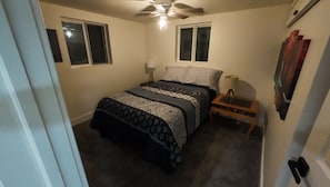 Ein Schlafzimmer mit einem Bett, zwei Fenstern, einem Deckenventilator, einem Nachttisch mit Lampe und einem Wandbild.