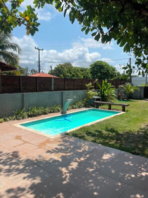 Una piscina rectangular con interior azul, rodeada por una valla de madera, un césped y una zona de patio con baldosas.