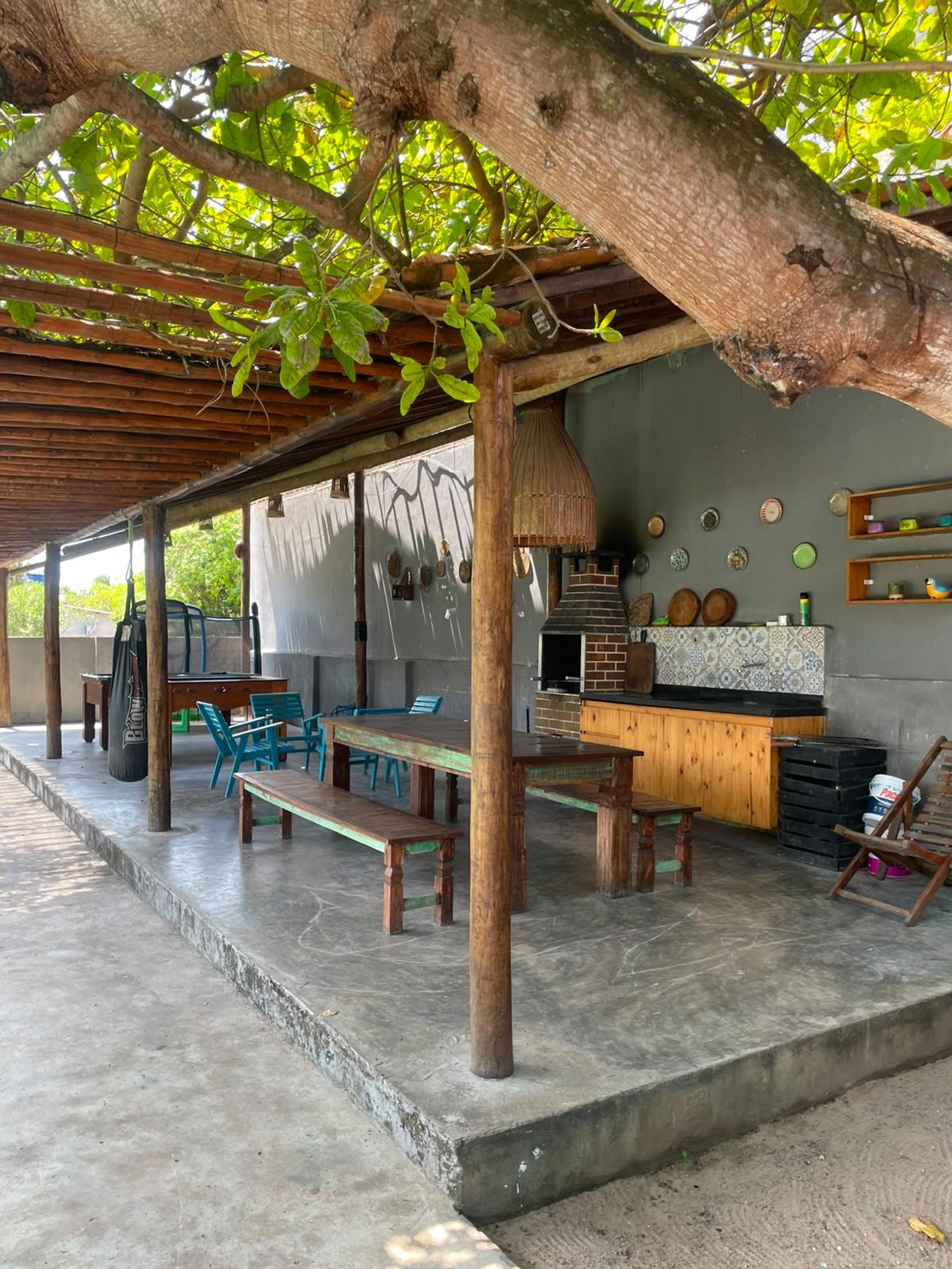 Patio esterno con pergola in legno, barbecue, tavolo da pranzo con sedie e angolo cottura con bancone e piatti decorativi.
