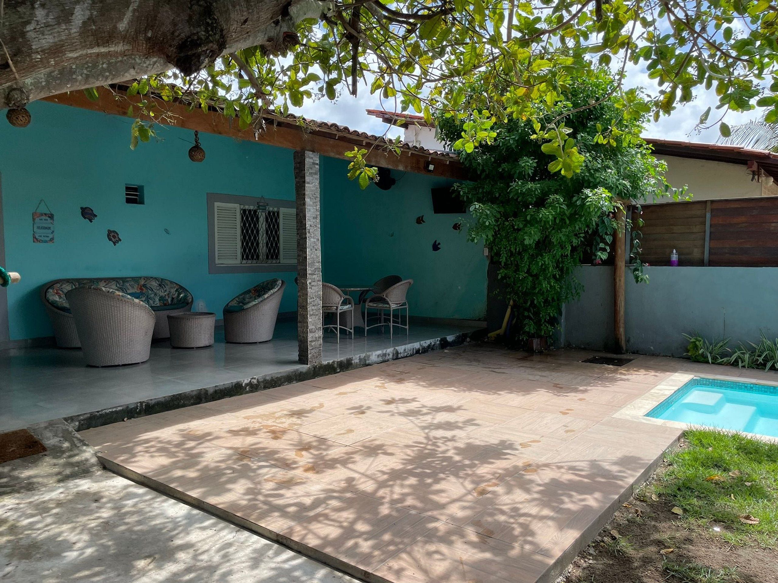 A covered outdoor area with a pool, wicker furniture, and a tree providing shade.