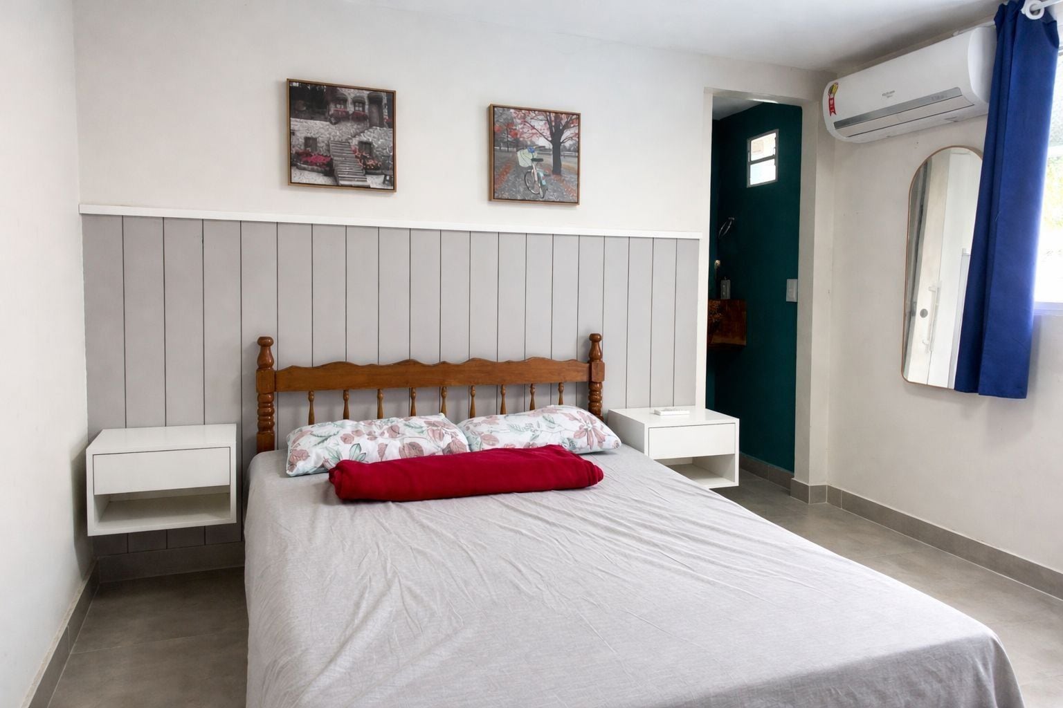 A bedroom with a wooden headboard, a bed with floral bedding, two bedside tables, and a wall-mounted air conditioning unit.