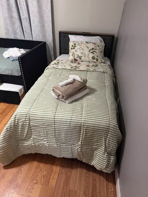A neatly made bed with striped bedding and a floral pillow, a folded towel on top, and a wooden floor.