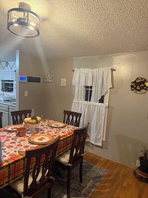 A dining area with a table set for four, chairs, a ceiling light fixture, a window with curtains, and a wall-mounted air conditioning unit.