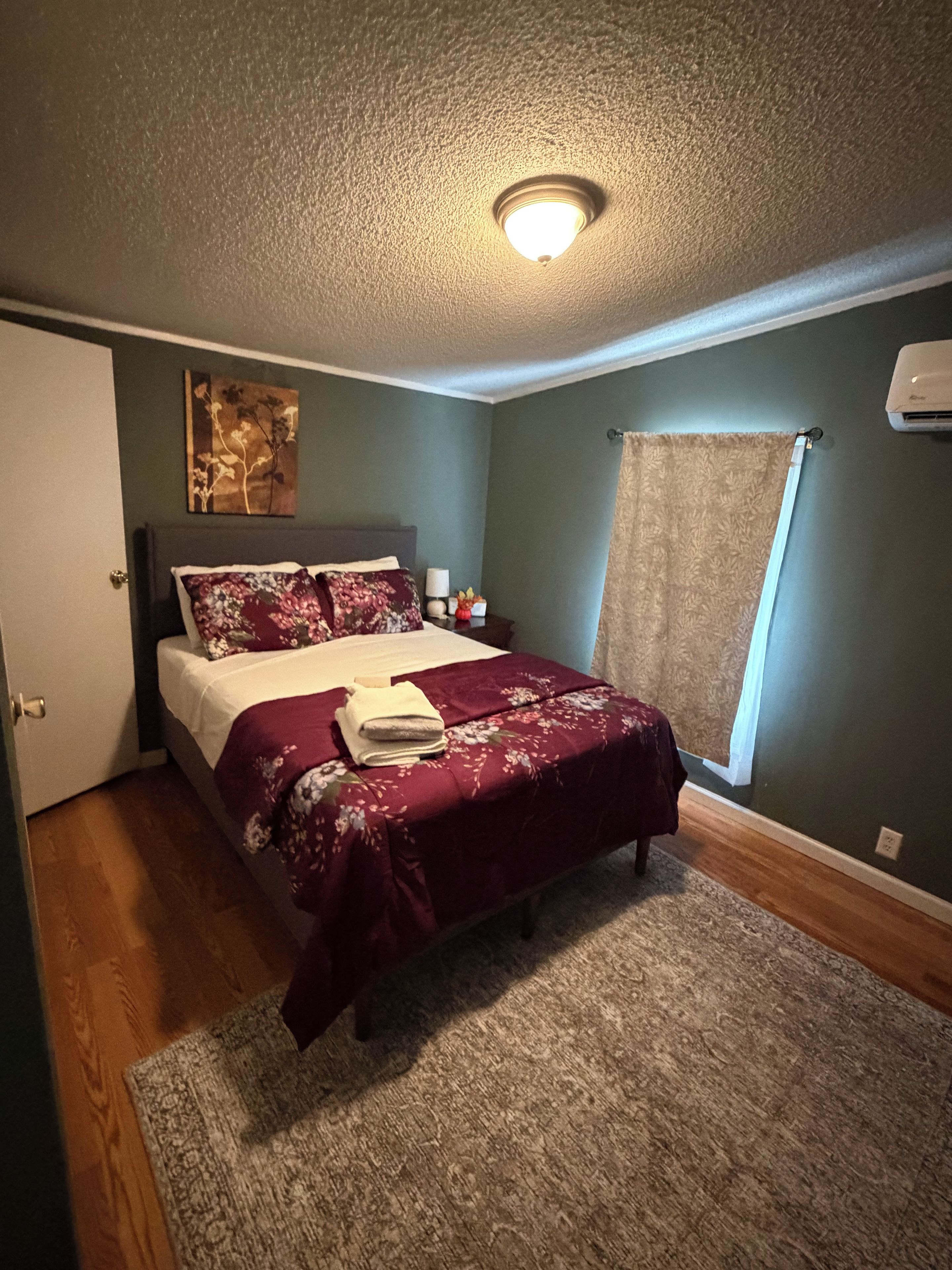 A bedroom with a bed, a nightstand, a wall-mounted air conditioner, and a window with a curtain.