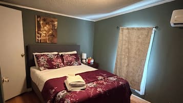 Une chambre à coucher comprenant un lit, une table de chevet, un climatiseur fixé au mur et une fenêtre avec un rideau.