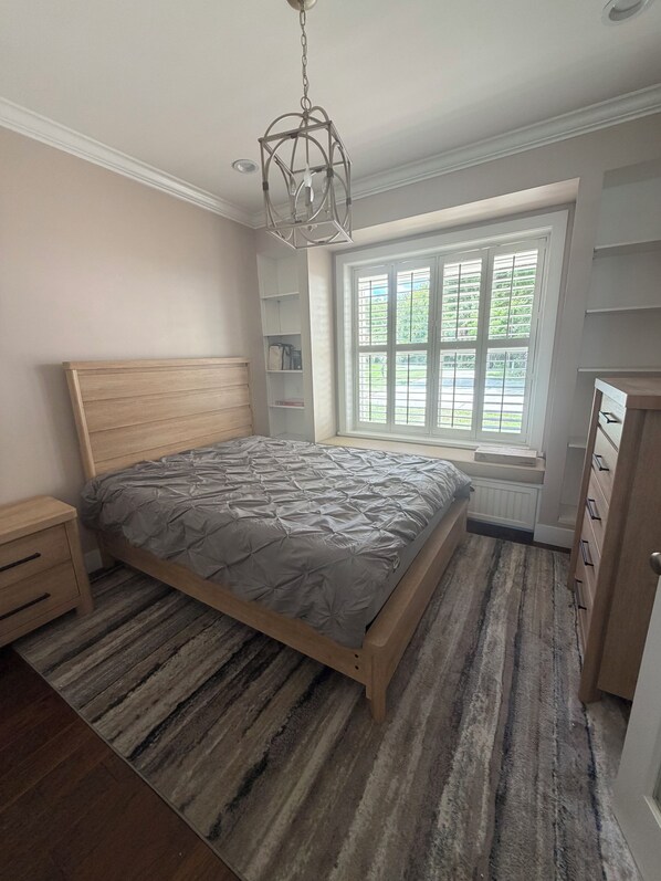 A bedroom with a wooden bed, a nightstand, a dresser, a window with blinds, and a hanging light fixture.