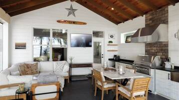A spacious living area with a white sofa, wooden dining table, and chairs. A kitchen with a brick backsplash and stainless steel appliances is visible.