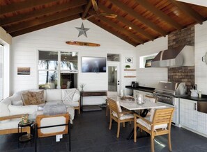 A spacious living area with a white sofa, wooden dining table, and chairs. A kitchen with a brick backsplash and stainless steel appliances is visible.