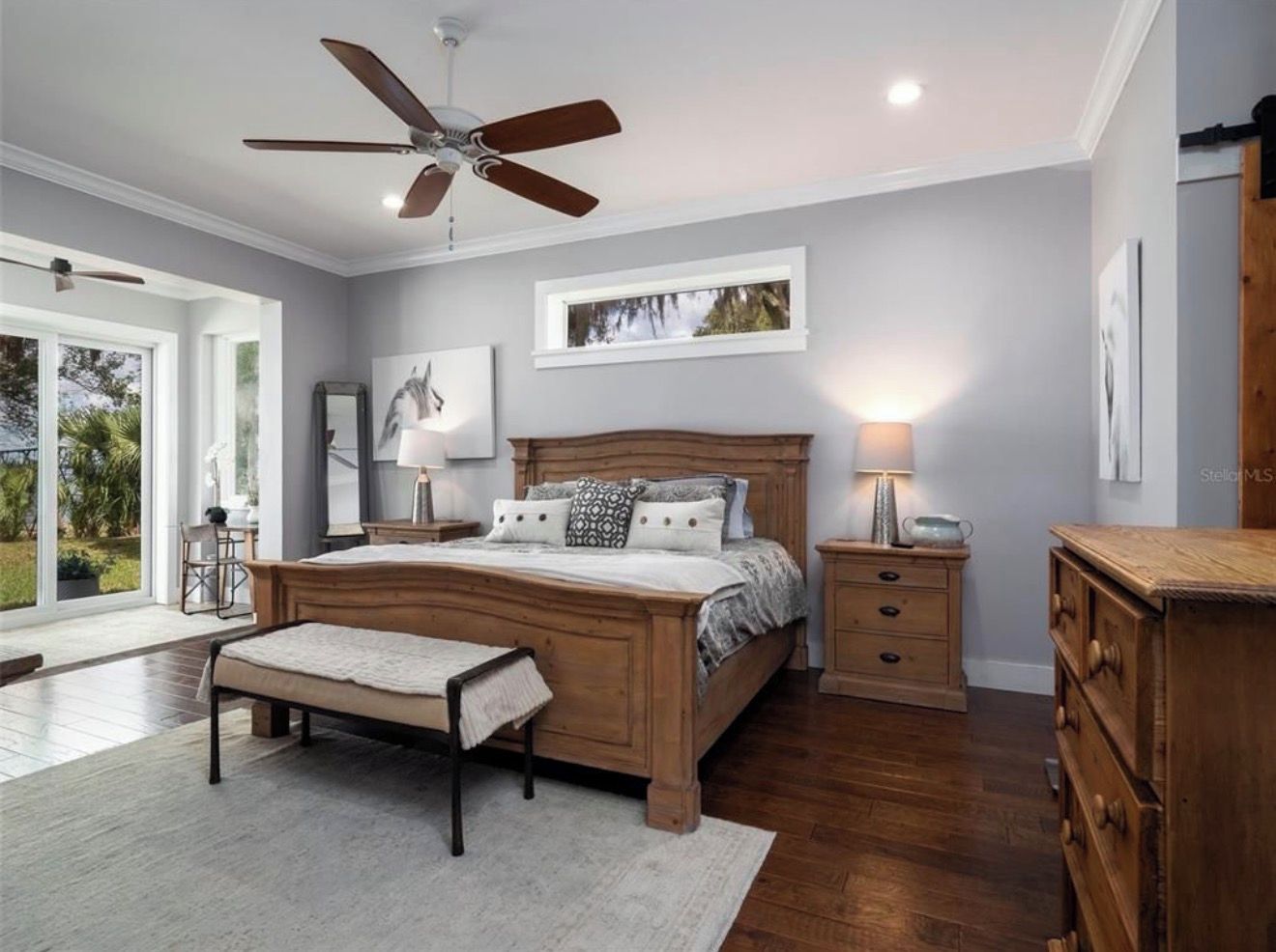 A bedroom with a wooden bed, bedside tables, a bench, a ceiling fan, and a large window.