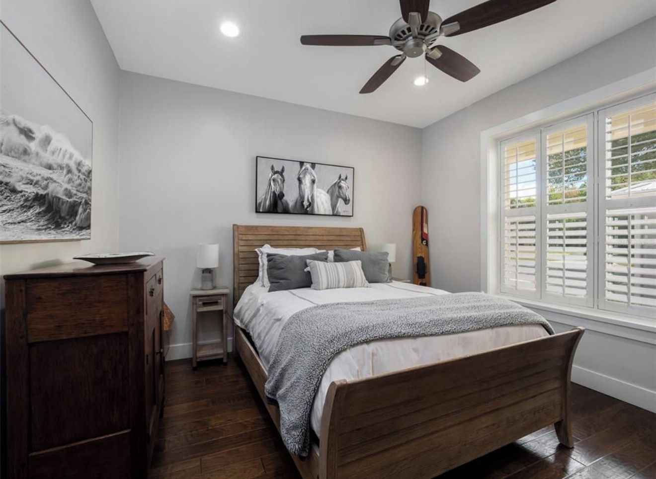 A bedroom with a wooden bed, a nightstand, a desk, a ceiling fan, and a window with blinds.