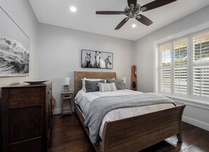Un dormitorio con cama de madera, mesita de noche, escritorio, ventilador de techo y ventana con persianas.