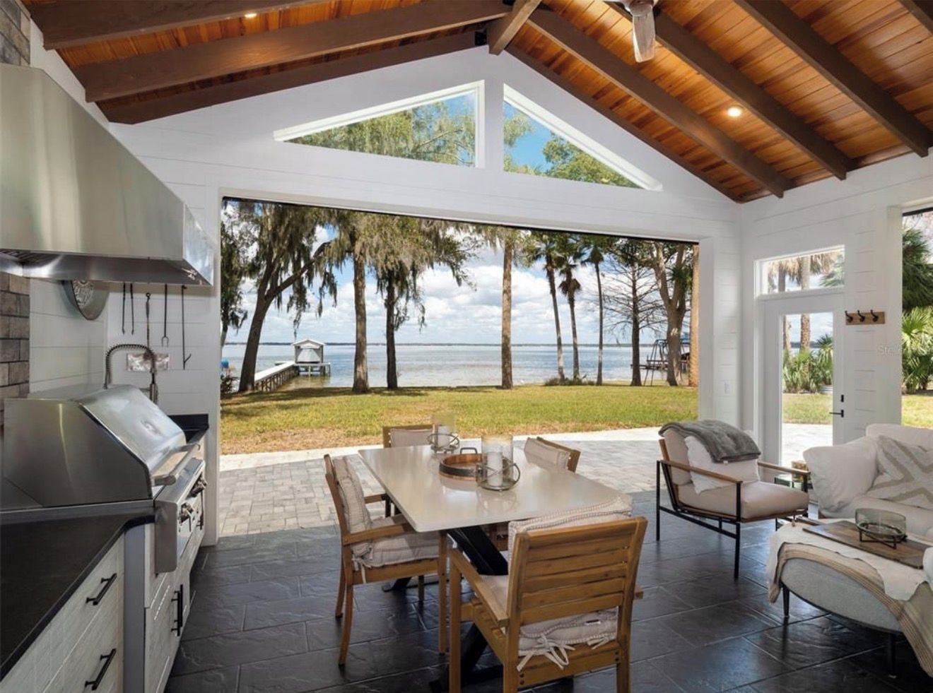 An outdoor kitchen with a grill, dining table, and chairs, overlooking a beach with palm trees and the sea.