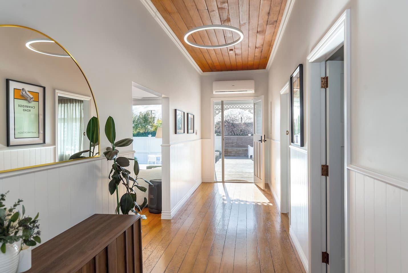 A hallway with wooden flooring, white walls, and a ceiling with wooden beams. There are framed pictures on the walls, a bench, and a potted plant.