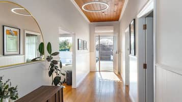 A hallway with wooden flooring, white walls, and a ceiling with wooden beams. There are framed pictures on the walls, a bench, and a potted plant.