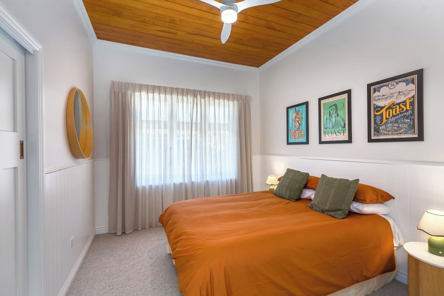 A bedroom with a large bed, a ceiling fan, a window with curtains, and three framed posters on the wall.