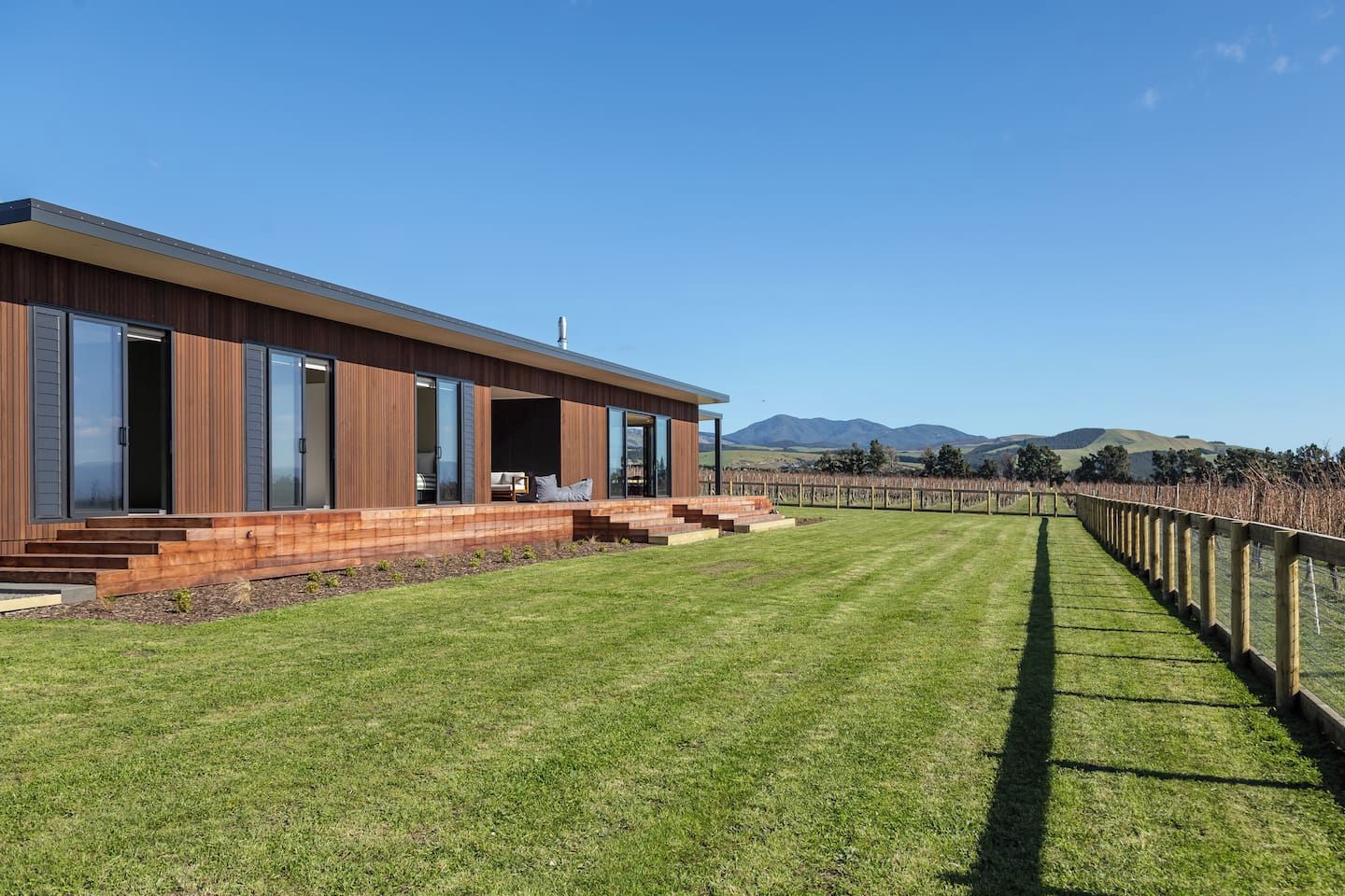A modern house with a wooden exterior, large windows, and a grassy lawn leading to a vineyard.