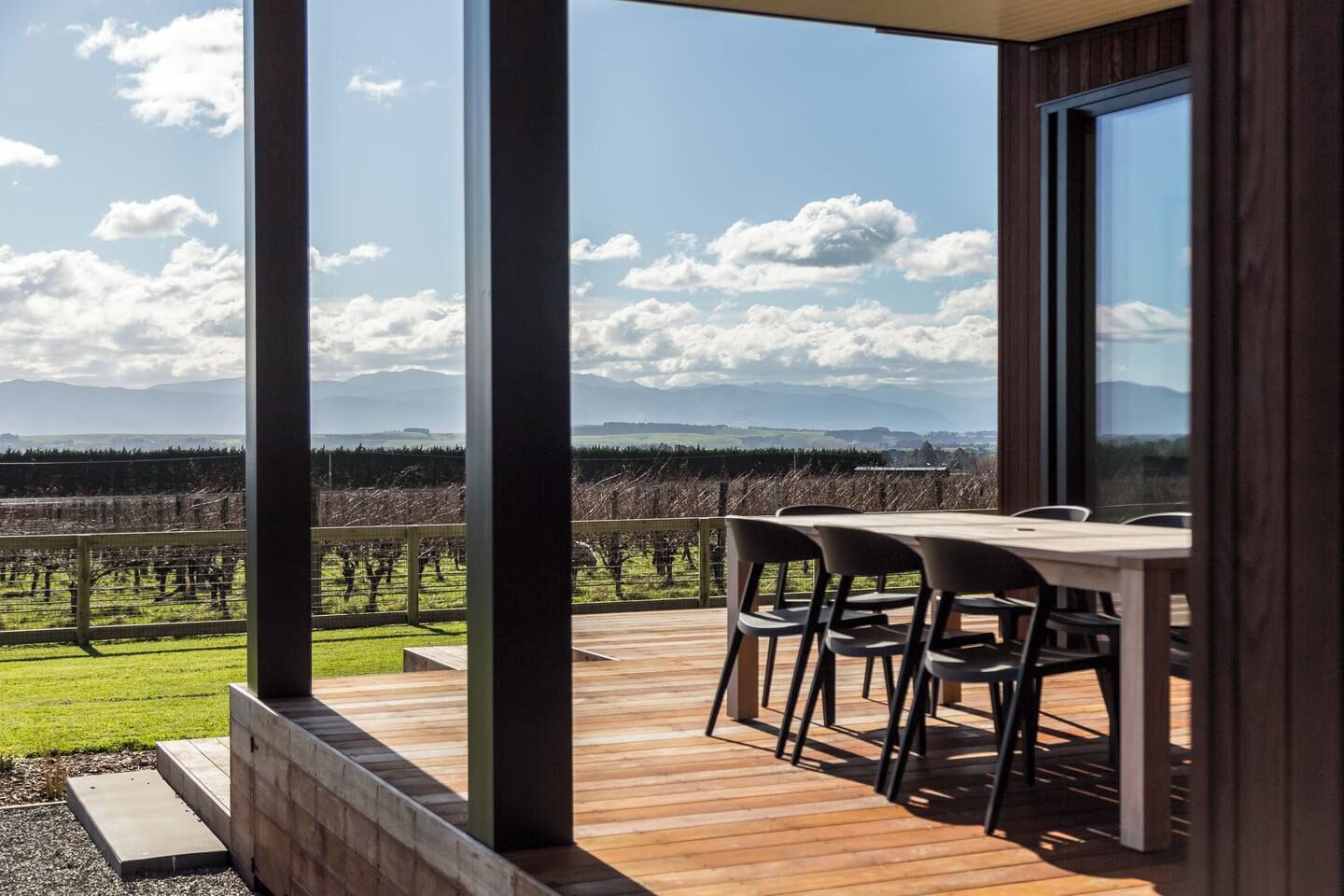 Een terras met een tafel en stoelen, met uitzicht op een wijngaard en verre bergen.