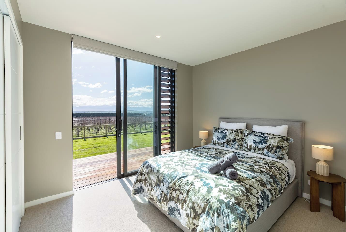 A bedroom with a large bed, a nightstand, a lamp, and a view of a vineyard through a sliding glass door.