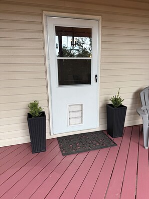 A white door with a window, two potted plants, a metal doormat, and a chair on a red wooden deck. - Lower Level Mountain Loft (Hot Springs)
