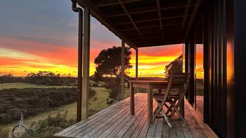 Une terrasse en bois avec une table et des chaises, offrant une vue sur un paysage arboré et un coucher de soleil.
