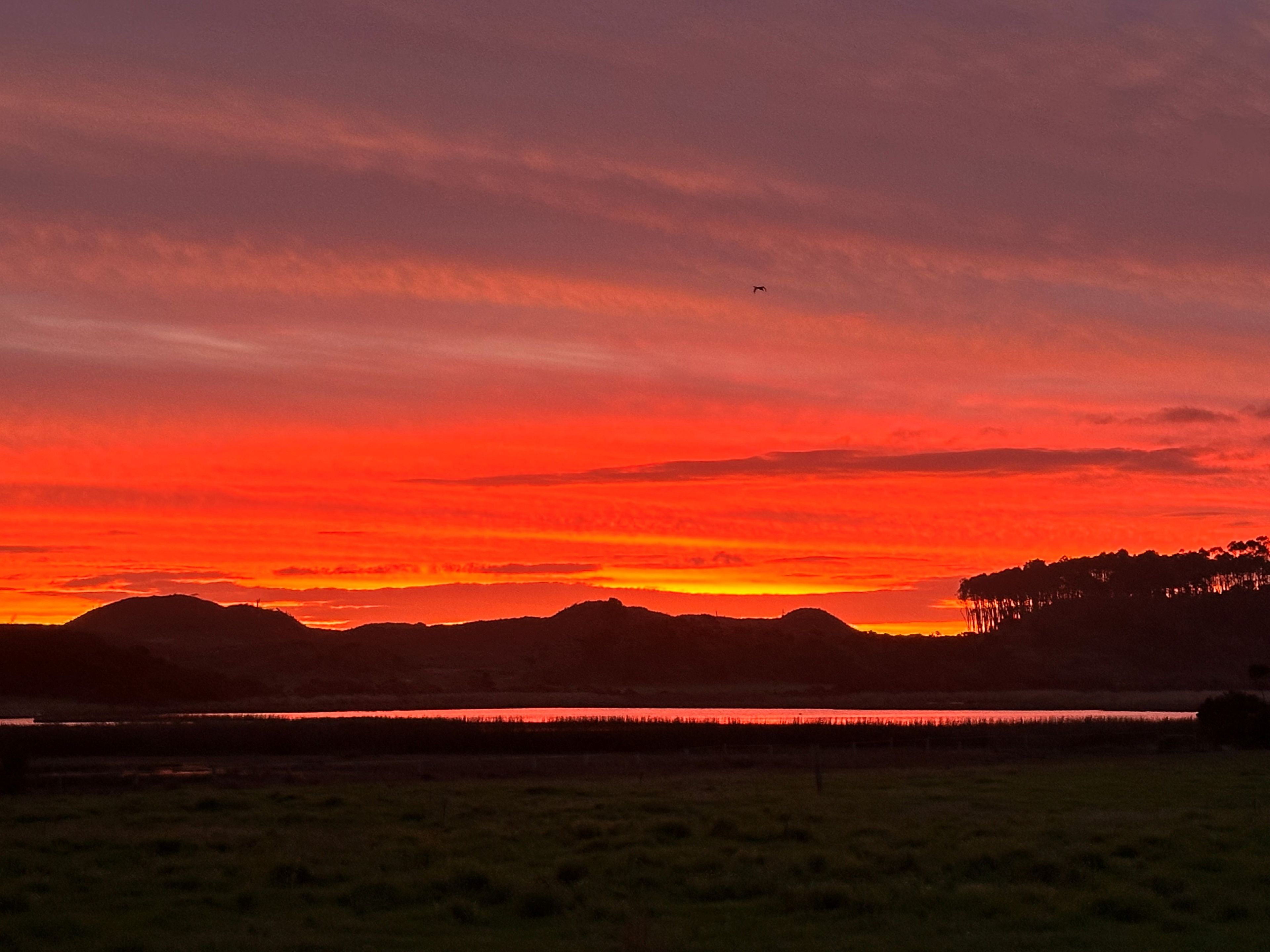 A sunset over a landscape with a body of water, hills, and a clear sky.