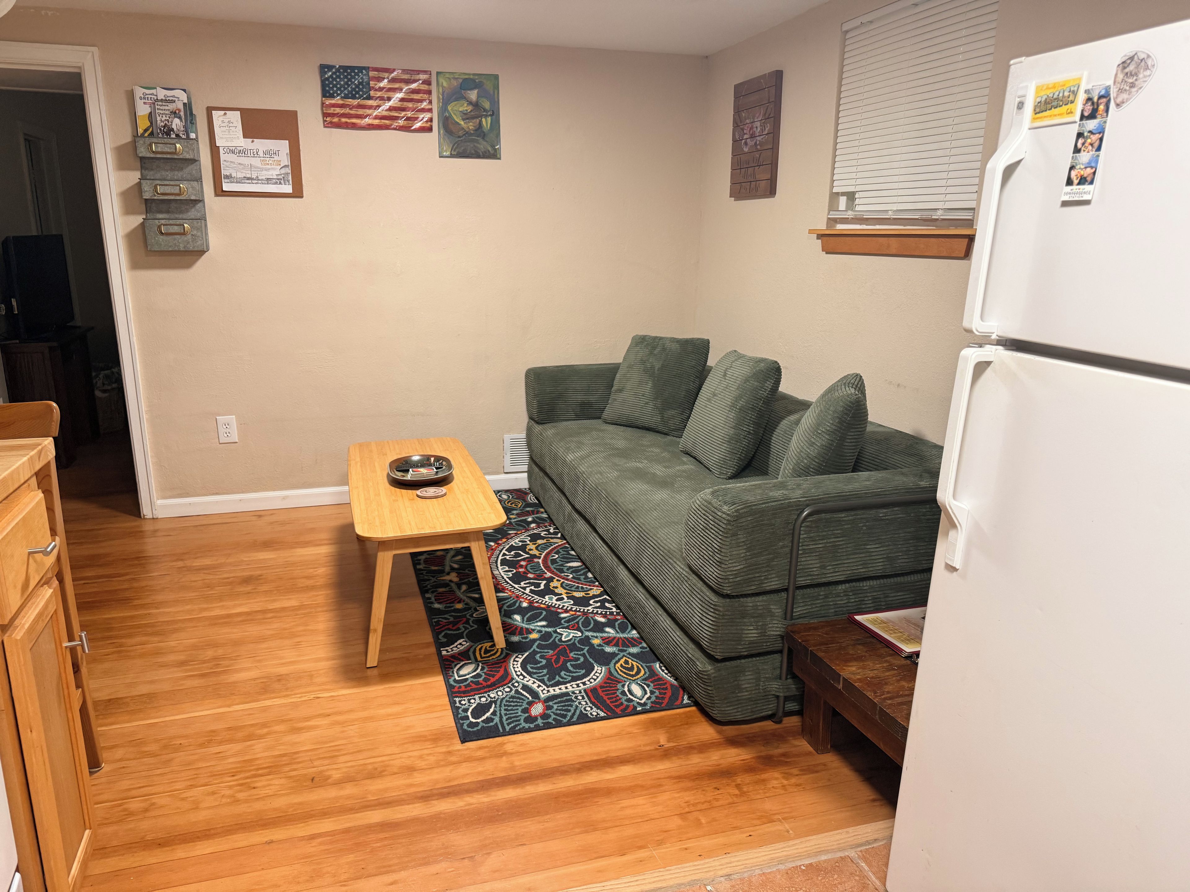 A compact living space with a green sofa, a small wooden table, a patterned rug, a refrigerator, and a bulletin board with various items pinned to it.
