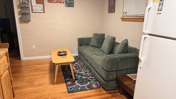 A compact living space with a green sofa, a small wooden table, a patterned rug, a refrigerator, and a bulletin board with various items pinned to it.