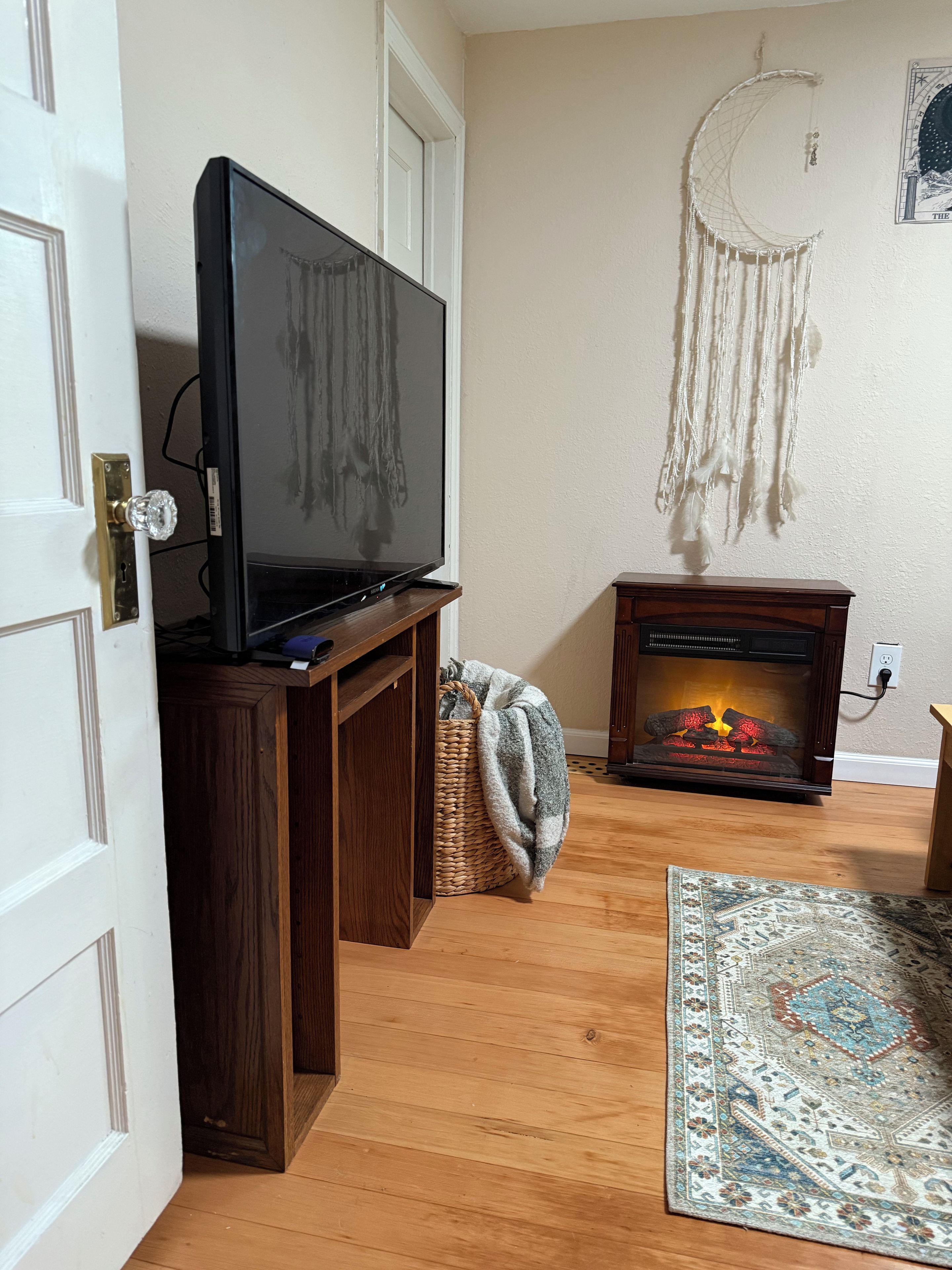 A living room with a television on a wooden stand, a fireplace with logs, a dreamcatcher on the wall, and a patterned rug on the floor.