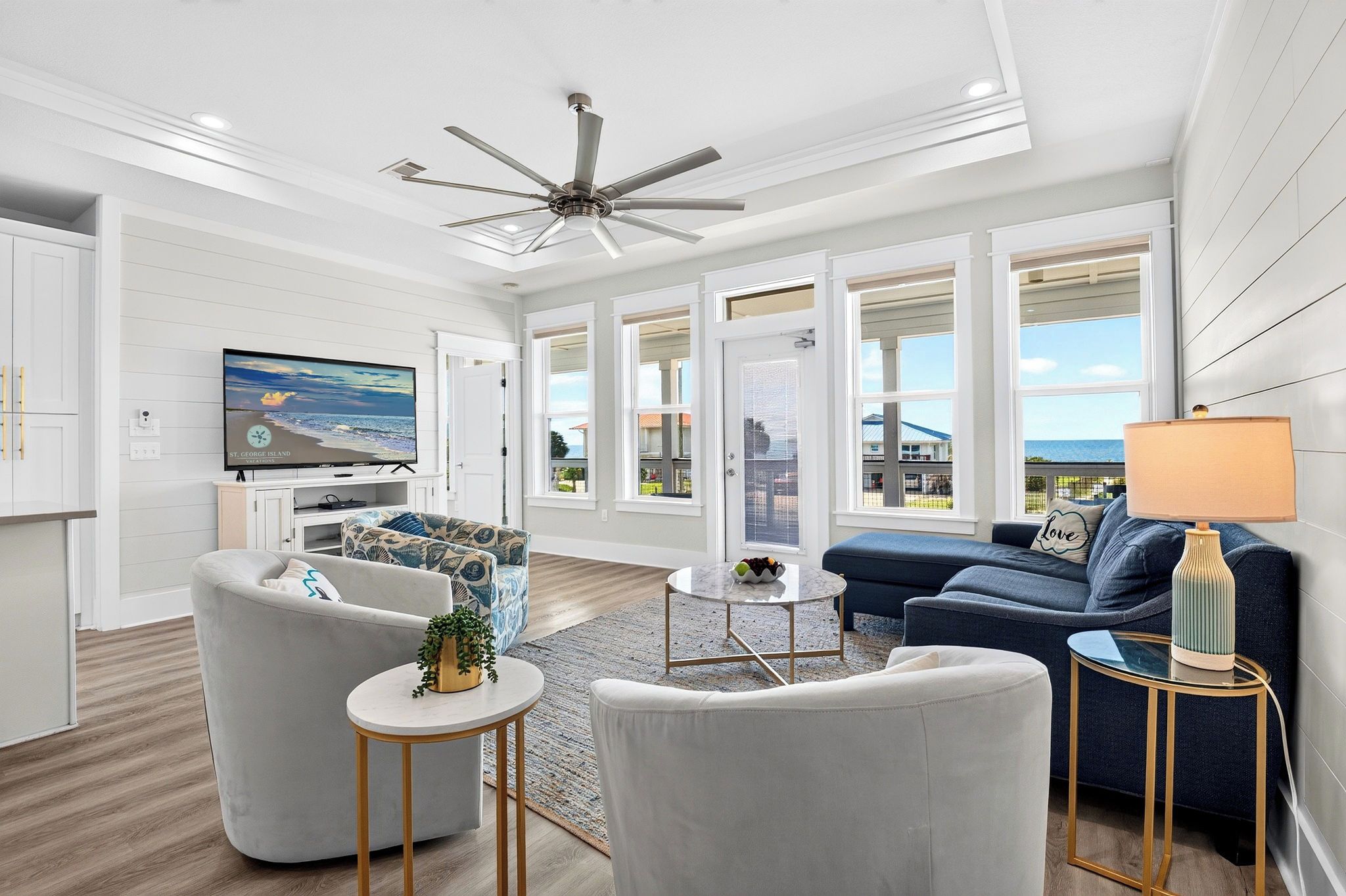 A modern living room with a flat-screen TV, comfortable seating, a ceiling fan, and large windows offering a view of the outside.