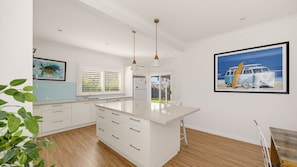 A modern kitchen with white cabinetry, a central island, pendant lighting, and a large framed picture of a van on the wall. - Rene's Cottage - a charming holiday rental nestled in the vibrant coastal town of Forster in NSW (Forster)