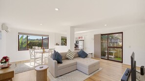 A modern living room with a sofa, a dining table, and chairs. There is a kitchen area visible through an open door. - Jasmine Retreat, nestled in the serene coastal neighbourhood of Black Head in NSW (Black Head)