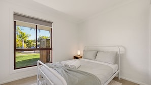 A bedroom with a bed, a nightstand, a lamp, and a window with a view of greenery. - Jasmine Retreat, nestled in the serene coastal neighbourhood of Black Head in NSW (Black Head)