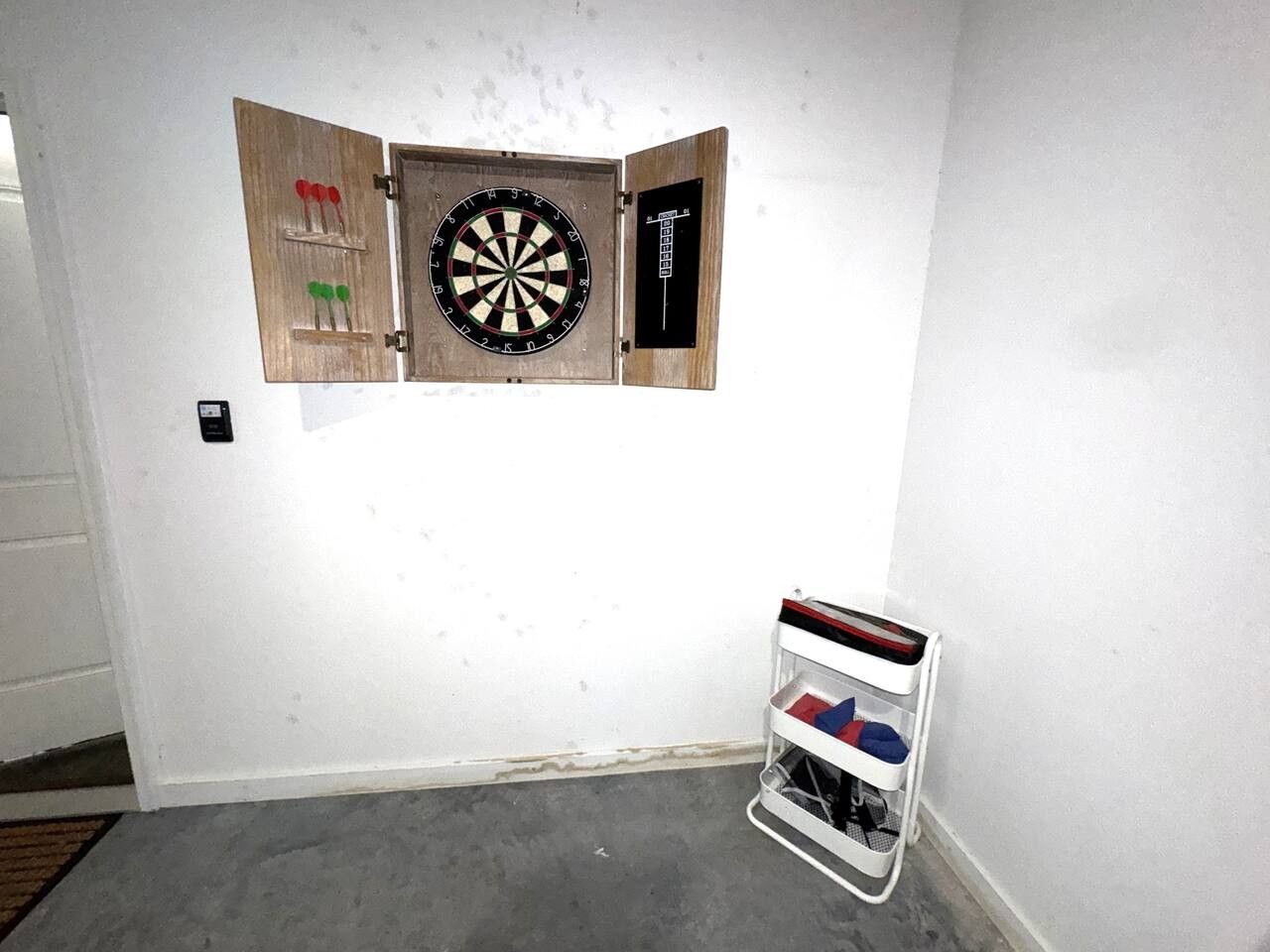 A dartboard mounted on a wall with a dartboard case and a small shelf with items.